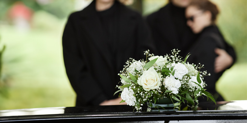 mourners in background with flowers on a coffin in the foreground