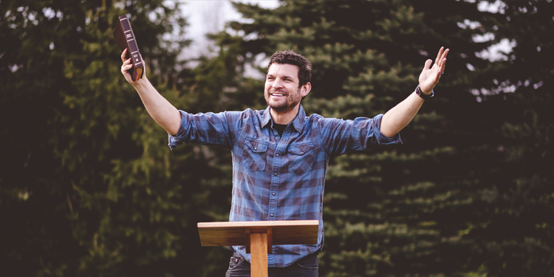 man preaching in front of pulpit outside with his hands in the air. He is holding a bible.